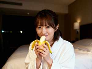 ホテルの部屋のベッドに座って顔の前にバナナを持っている若い女性を、正面カメラのアング...。