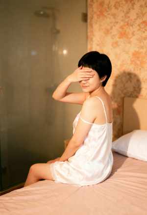白いシルクのナイトガウンを着て手で目を覆い、ベッドに座っている若い女性を横から撮った...。