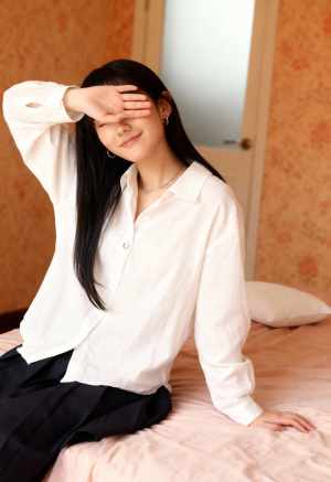 白いボタンダウンシャツと黒いスカートを着て、手で目を覆い、ベッドに座っている若い女性...。