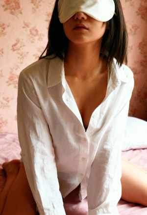 白いシャツを着て目隠しをしてベッドに座る女性を横顔カメラのアングルから写実的に撮影したもの。
