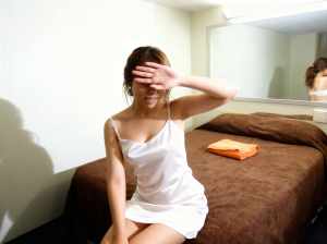 寝室のベッドに座り、手で目を覆っている若い女性を横から撮った写実的な写真。