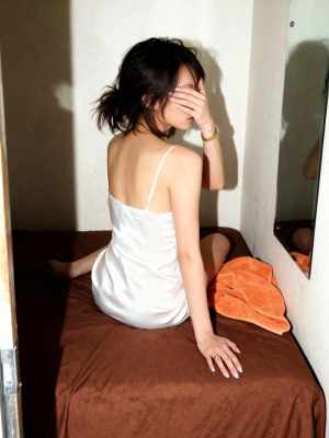 白いスリップドレスを着て手で顔を覆い、ベッドに座っている若い女性を横から撮った写実的な写真。