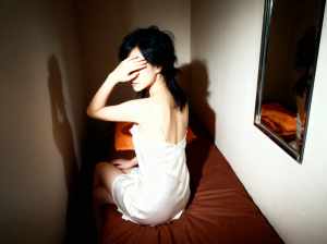 白いスリップドレスを着て、薄暗い部屋のベッドに一人で座っている若い女性を横から撮った...。