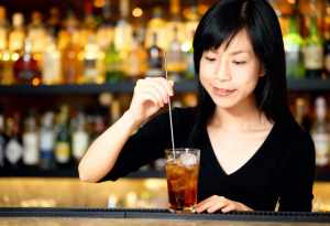 黒いVネックのシャツを着た長い黒髪の若いアジア人女性が、酒瓶の棚を背景にした薄暗いバ...。