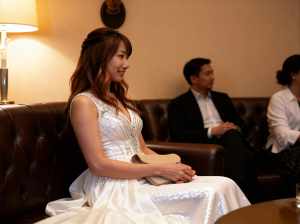 白いウェディングドレスを着た女性が薄暗い部屋で茶色の革張りのソファに座り、笑顔で横を...。