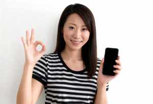 無地の白い背景の前に立ち、右手に黒いスマートフォンを持ち、左手で OK サインを出し...。