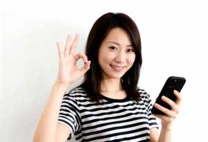 無地の白い背景の前に立ち、右手に黒いスマートフォンを持ち、左手で「OK」のジェスチャ...。