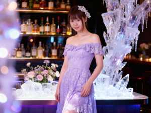 ラベンダーのレースのドレスを着た若い女性がボトルを背景にバーの前に立っている様子を、...。