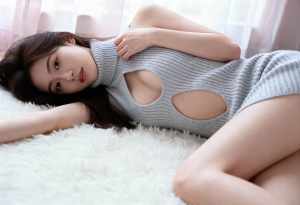 カットアウトのあるグレーのニットドレスを着て、リラックスしたポーズでふわふわの白い敷...。