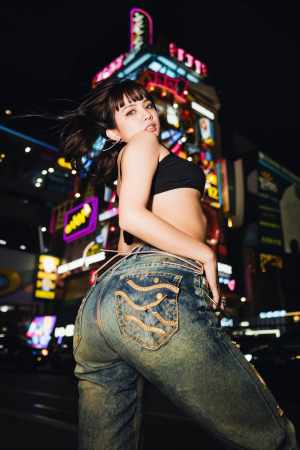 夜のネオン輝く街並みの中で黒いクロップトップとジーンズを着てポーズをとる女性を後ろか...。