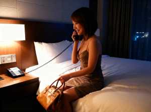 ホテルの部屋のベッドに座り、茶色のハンドバッグを持ちながら電話で話している女性。