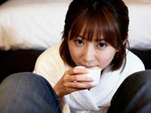 ソファに座り、手に白いカップを持ち、瞑想的な表情でカメラを直接見つめている若い女性の接写。