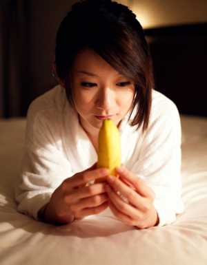 ベッドに座って顔の前に黄色いバナナを持ち、瞑想的な表情でそれをじっと見つめている若い...。