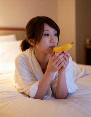 白いバスローブを着た若い女性がベッドに横たわり、顔の前にバナナを持っている様子を横か...。