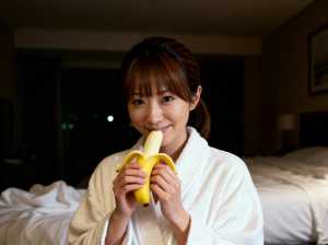 白いローブを着た若い女性がベッドに座り、口の前にバナナを持ってカメラに向かって微笑ん...。