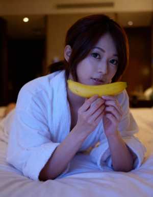 白いバスローブを着た若い女性がベッドに横たわり、顔の前にバナナを持ち、無表情でカメラ...。