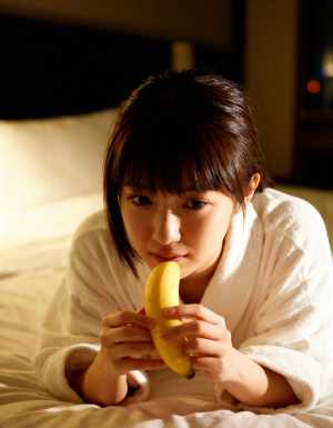 ベッドに座ってバナナを持ち、無表情でカメラを見つめる若い女性を正面から写実的に撮影したもの。