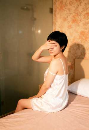 白いシルクのナイトガウンを着て手で目を覆い、ベッドに座っている若い女性を横から撮った...。