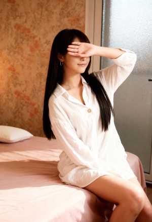 白いシャツワンピースを着て右手で目を覆い、ベッドに座る若い女性を横から写実的に撮影した写真。