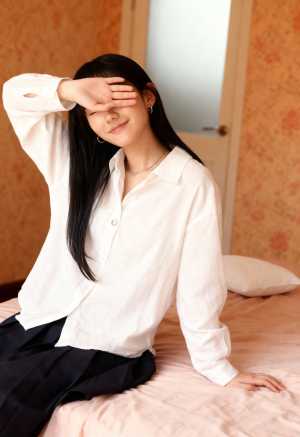 白いボタンダウンシャツと黒いスカートを着て、手で目を覆い、ベッドに座っている若い女性...。