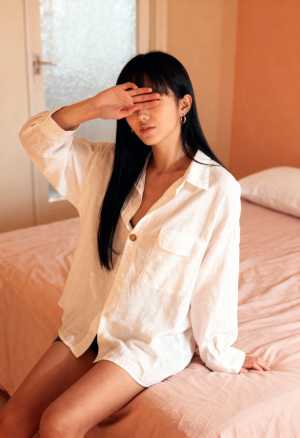 白いボタンダウンシャツと黒いショートパンツを着て、手で目を覆い、ベッドに座っている若...。