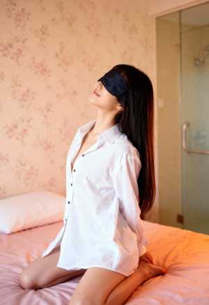 白いボタンダウンのシャツを着て、目隠しをしてベッドに座っている若い女性を横から撮った...。