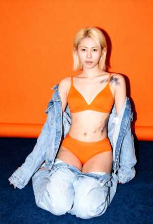 オレンジ色の背景の前で床に座り、オレンジ色のブラジャーとそれに合わせたオレンジ色のパ...。