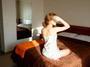 ミニマルな装飾と壁に大きな鏡のあるモダンな寝室で、白いナイトガウンを着てベッドに座っ...。