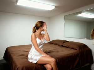 薄暗い寝室のベッドに座り、手で顔を覆っている若い女性を横から写実的に撮影した写真。