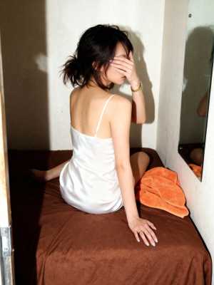 白いスリップドレスを着て手で顔を覆い、ベッドに座っている若い女性を横から撮った写実的な写真。