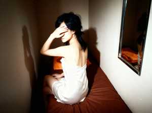 白いスリップドレスを着て、薄暗い部屋のベッドに一人で座っている若い女性を横から撮った...。