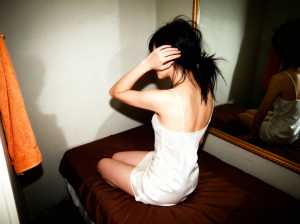 薄暗い部屋でベッドに座り、白いスリップドレスを着た若い女性を横から撮った写実的な写真。