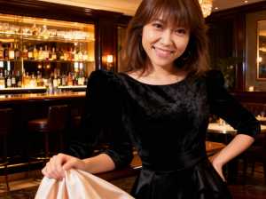 薄暗いバーに立ち、布を手に持ちながらカメラに向かって微笑む若い女性のポートレート。