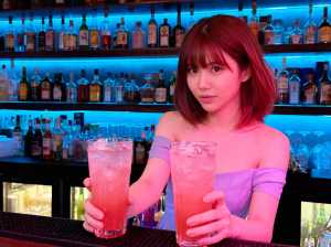 短い赤い髪の若い女性がバーカウンターの後ろに立ち、ピンク色の飲み物が入った背の高いグ...。