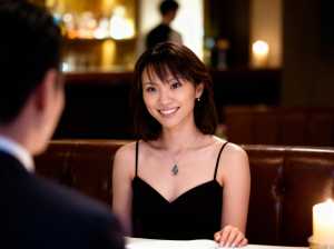 黒いドレスと銀のネックレスを身に着け、レストランのテーブルに座り、後ろの男性と会話し...。