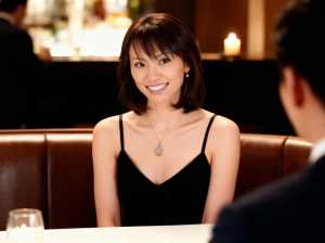 黒いドレスを着て、涙の形のペンダントが付いたネックレスを着て、レストランのテーブルに...。