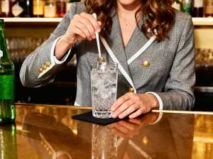 バーに座り、氷とスプーンを入れた水の入ったグラスを持っている、グレーのチェック柄のブ...。