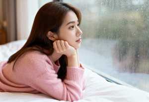 ピンクのセーターを着た長い茶色の髪の若い女性が、曇りガラスの窓の前のベッドに横たわり...。