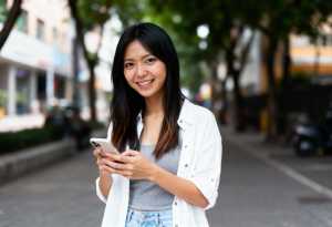 街の通りの真ん中に立ち、スマートフォンを手に持ち、カメラに向かって微笑むアジア系の若い女性。