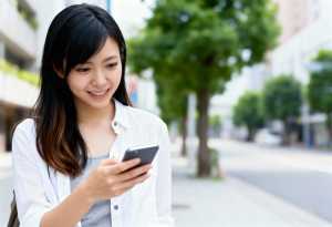 都会のベンチに座り、笑顔で携帯電話を見ているアジア系の若い女性。