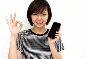 画像の中央に立ち、右手に黒いスマートフォンを持ち、左手でOKサインを出している若い日...。