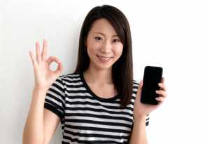 無地の白い背景の前に立ち、右手に黒いスマートフォンを持ち、左手で OK サインを出し...。