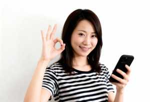 無地の白い背景の前に立ち、右手に黒いスマートフォンを持ち、左手で「OK」のジェスチャ...。