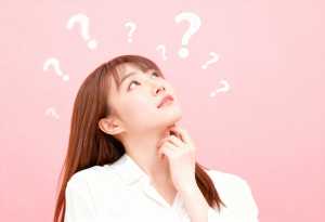 柔らかいピンクの背景に、白いブラウスを着た長い茶色の髪の若い女性が、頭上に疑問符を浮...。