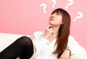 ピンクの背景に白いソファに座り、頭の上に疑問符が浮かび、思慮深い表情で見上げる若い女性。