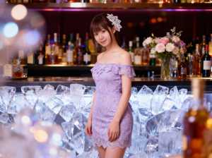 薄紫のレースのドレスを着て、ボトルと氷がいっぱいのバーの前に立つ若い女性を正面から写...。