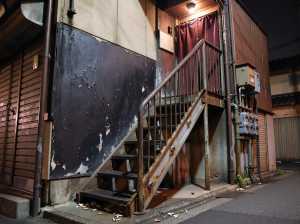 ローアングルから写実的に撮影したもので、夜の薄暗い路地と、錆びた金属製の階段が、塗装...。