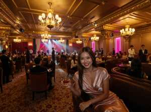 高級レストランに座って赤ワインのグラスを持ち、カメラに向かって微笑む女性を横から写実...。