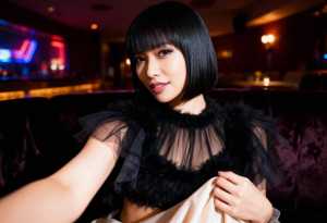 薄暗いレストランのソファに座るボブヘアの若い女性が、エッジのフリルが付いた薄手の黒い...。