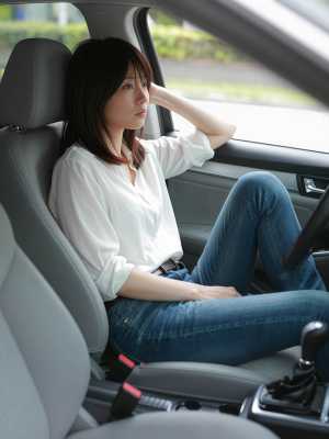 白いブラウスとブルー ジーンズを着て、現代の車の運転席に座っている若いアジア人女性を...。